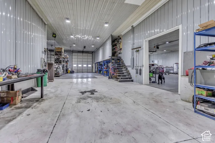 Spacious interior featuring a large overhead door, concrete flooring, corrugated metal walls, exposed overhead lighting, and an integrated metal staircase
