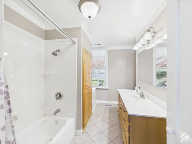 Built-in bathtub with shower, wood-finish vanity with dual sinks, overhead vanity lighting, tiled flooring, and built-in wood-finish cabinetry