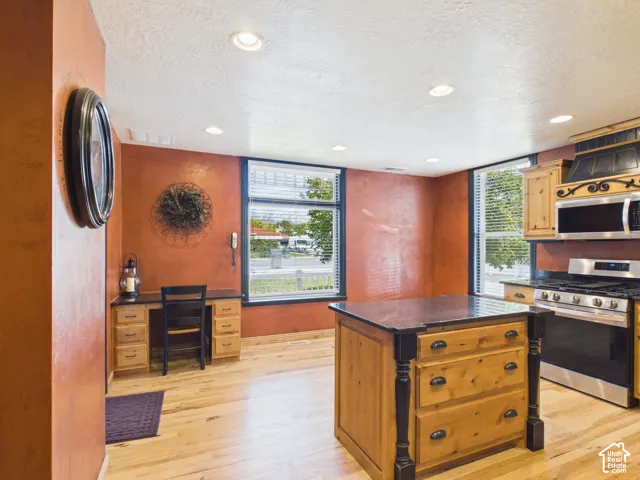 Light wood-finish flooring, recessed lighting, built-in wooden desk, kitchen island with dark countertop, stainless steel range and microwave