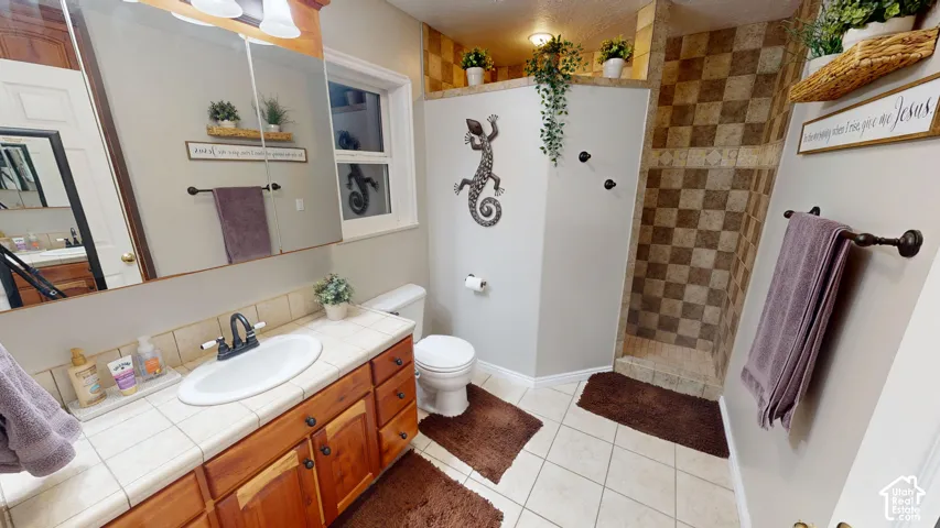Ceramic tile flooring, wood-finish vanity with ample storage, tiled countertop with integrated sink, walk-in shower with checkered tile, and recessed lighting