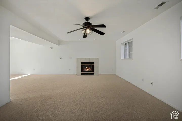 Spacious room featuring neutral carpeting, a central ceiling fan with integrated lighting, a fireplace with a light-toned tile surround, and a window providing natural illumination