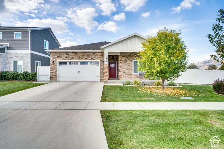 Stone veneer facade, attached two-car garage, concrete driveway, front yard with mature landscaping, and white privacy fencing