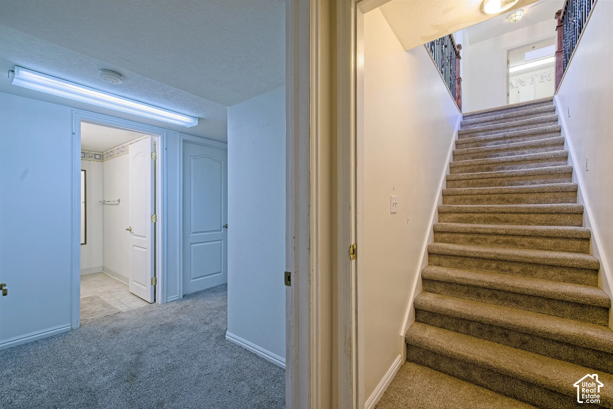 Carpeted staircase with an ornamental metal handrail, recessed wall paneling, and an overhead light fixture