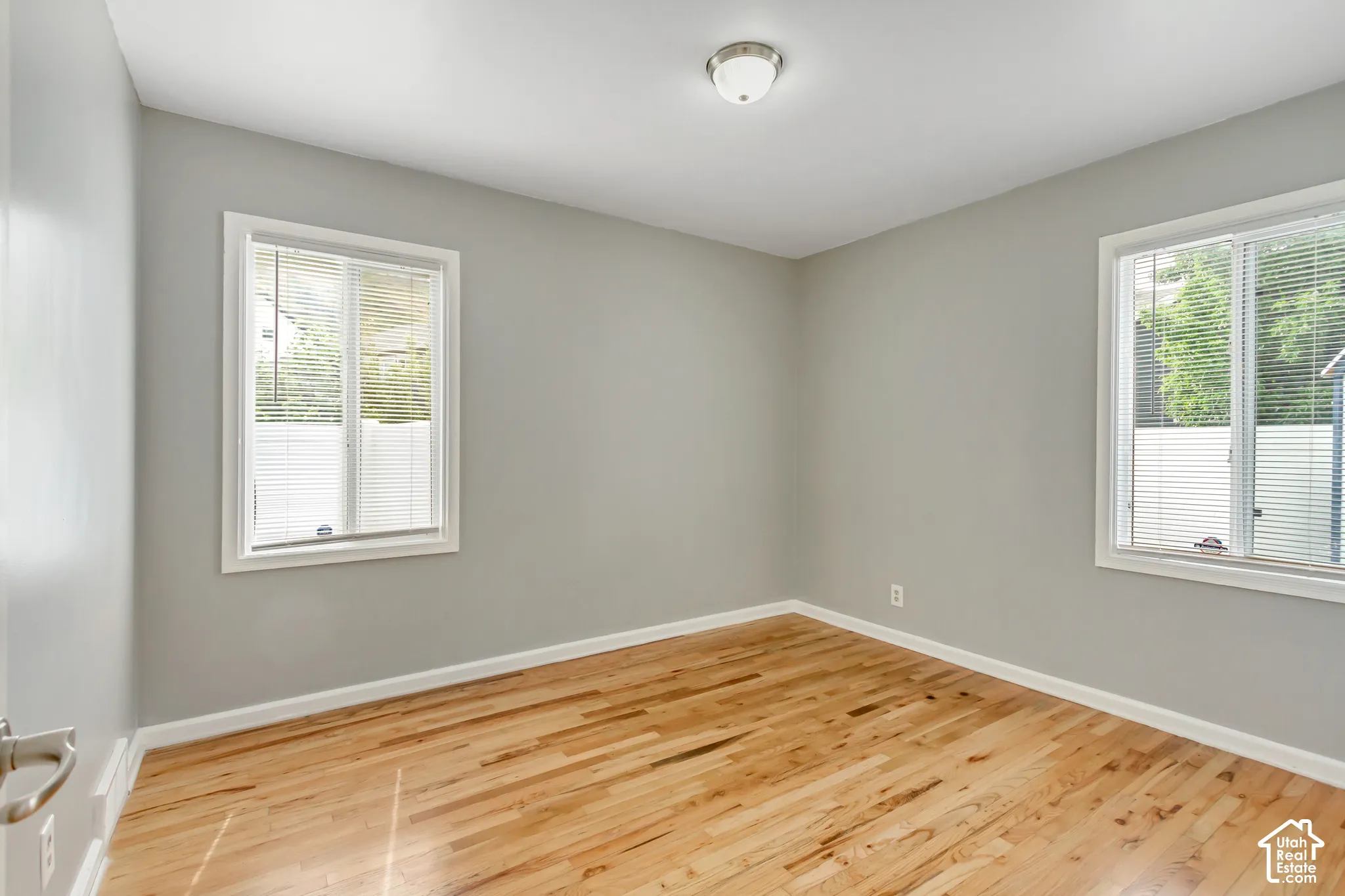 Light wood-finish flooring, two windows with white blinds, and a semi-flush mount ceiling light fixture