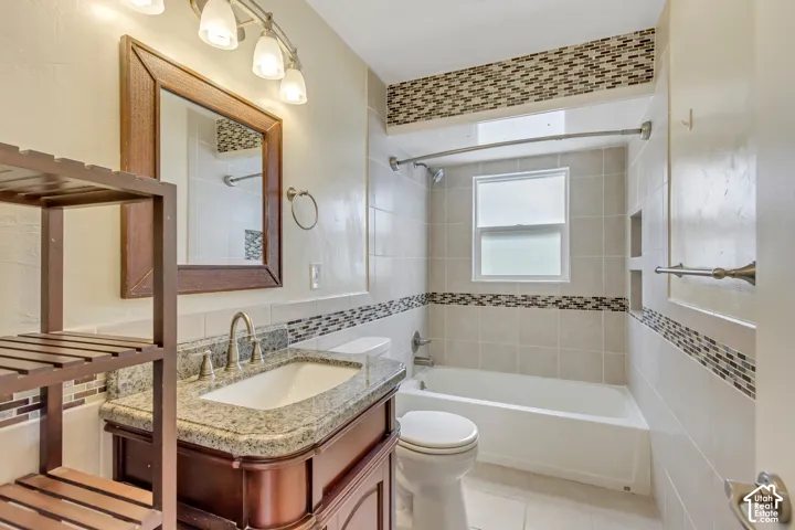Vanity with granite countertop and undermount sink, tiled bathtub surround with mosaic accent, built-in shelving in shower, and light fixture with multiple frosted glass shades