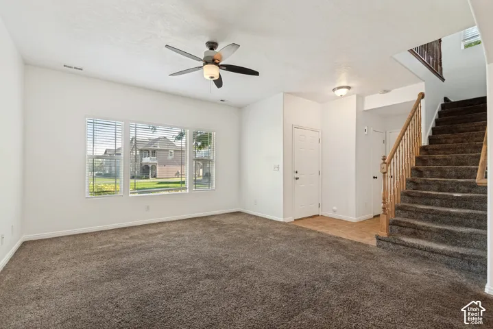 Carpeted living area featuring a ceiling fan with integrated lighting, three windows with blinds, and a staircase with a wood-finish railing