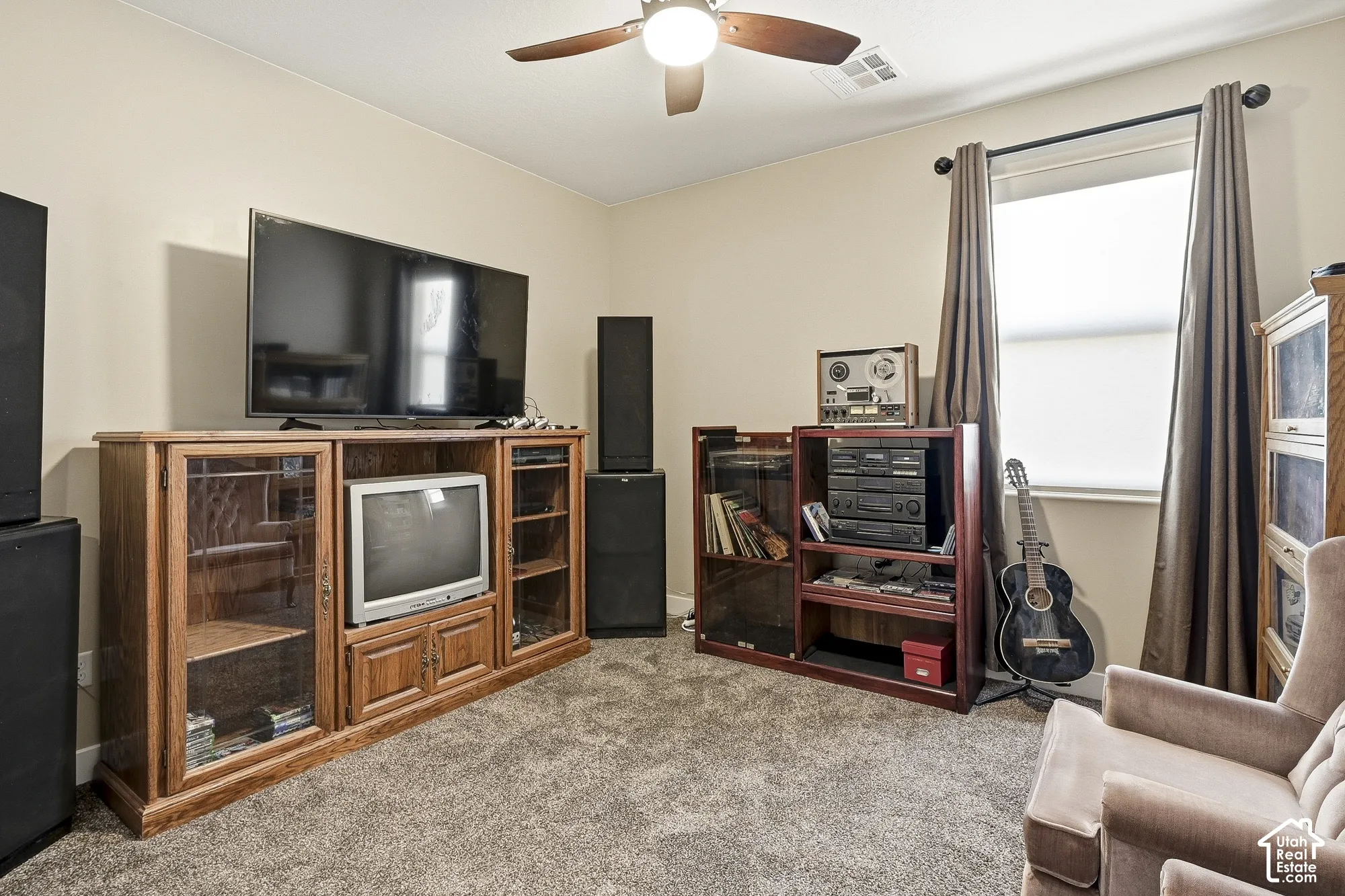 Textured carpet flooring, ceiling fan with light fixture, window with roller shade and curtains, and neutral wall paint