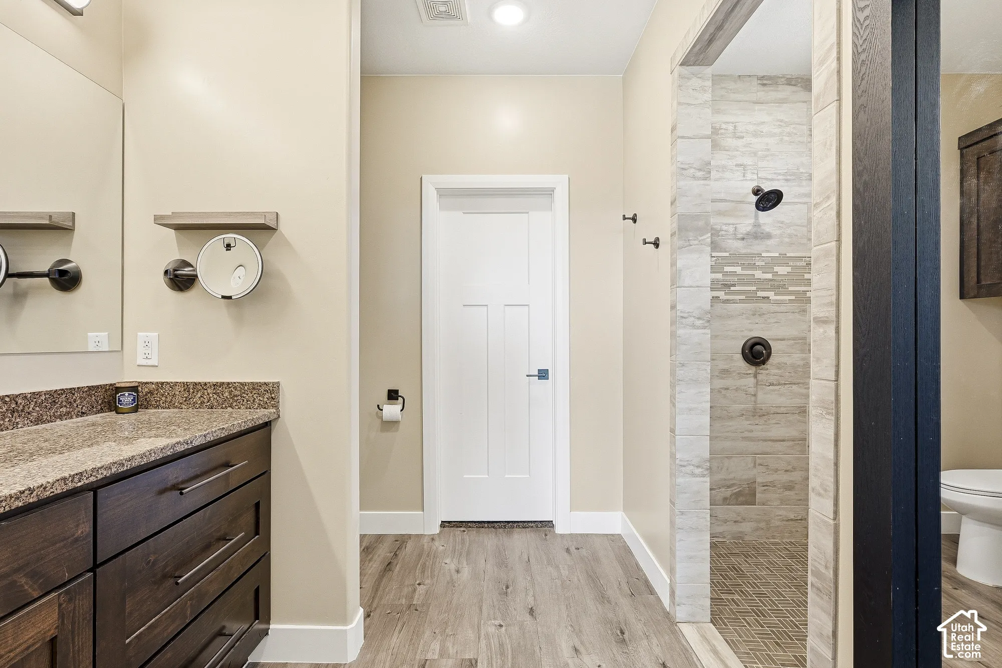 Wood-finish flooring, dark wood-tone vanity with stone countertop, large mirror, and tiled walk-in shower with decorative mosaic band