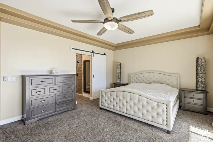 Tray ceiling with crown molding, ceiling fan with light fixture, carpeted flooring, and barn door entry