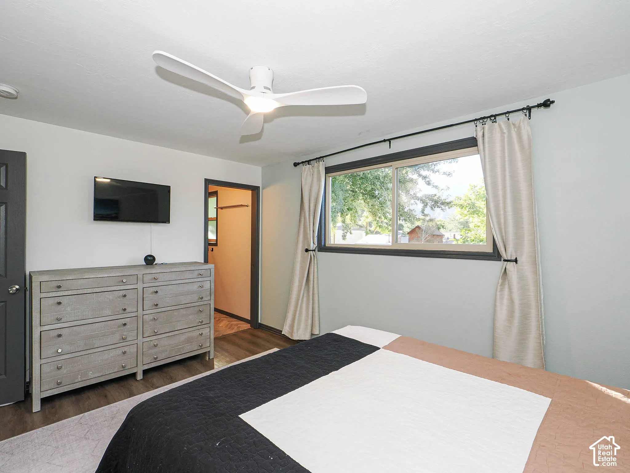 Bright secondary bedroom featuring hardwood flooring, a ceiling fan with integrated lighting, large window with natural light and custom drapery, and direct access to an ensuite bathroom