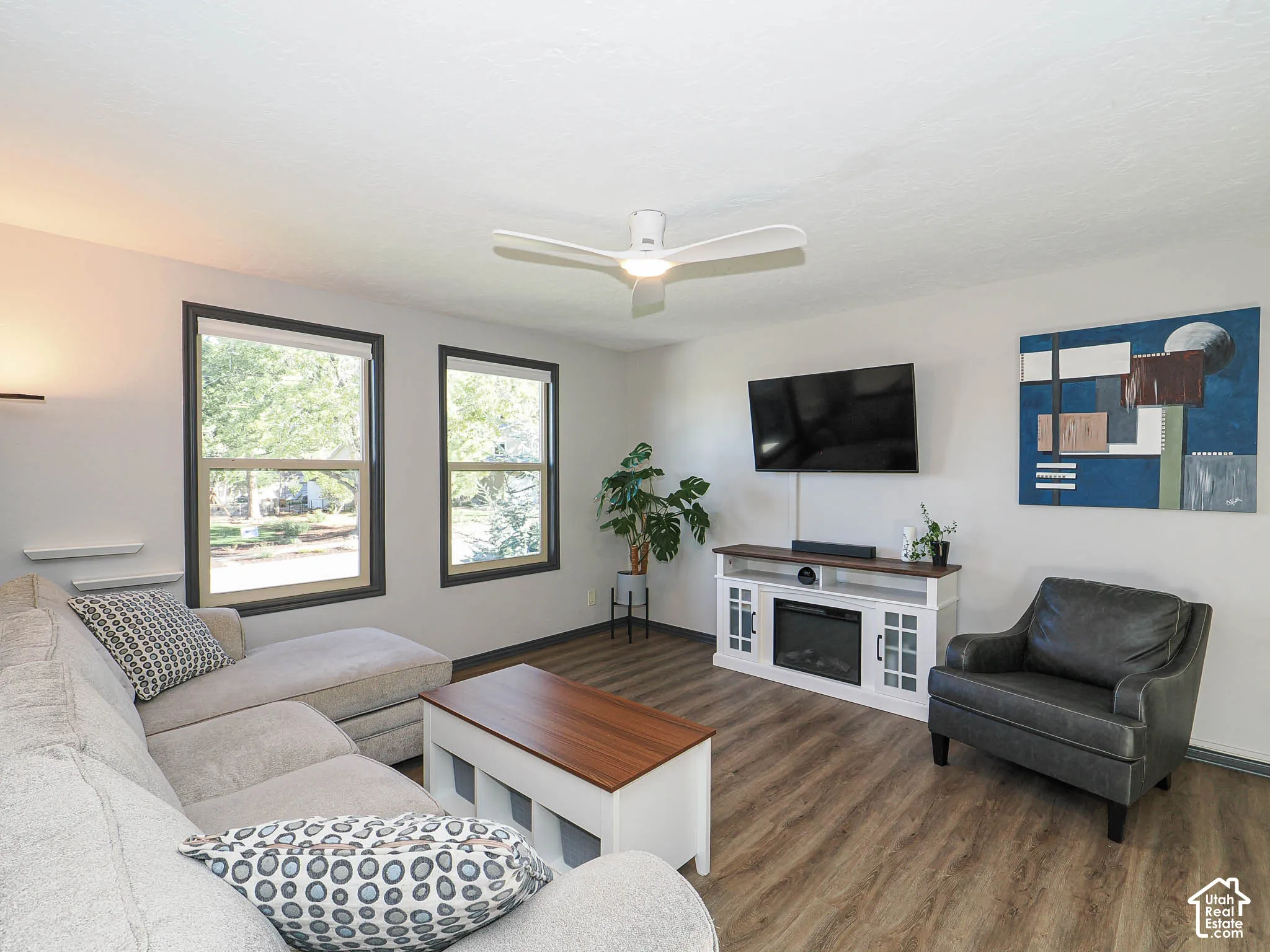 Wood-finish flooring, two large windows, ceiling fan, and built-in media console with integrated electric fireplace