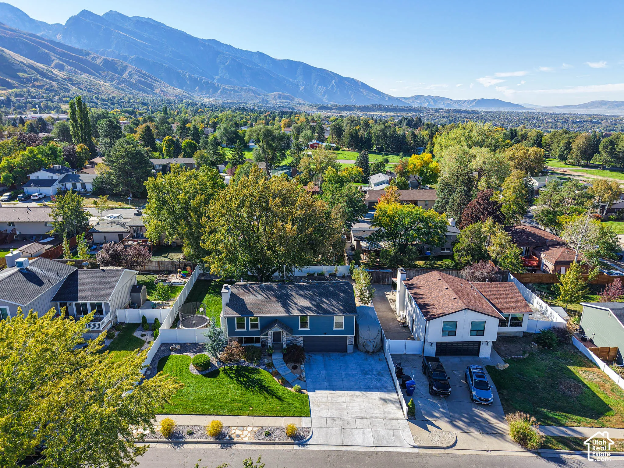 Striking blue contemporary home featuring a spacious driveway with multiple parking spaces, mature landscaping with established trees throughout the grounds, manicured lawn, and mountain backdrop