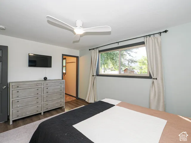 Bright secondary bedroom featuring hardwood flooring, a ceiling fan with integrated lighting, large window with natural light and custom drapery, and direct access to an ensuite bathroom