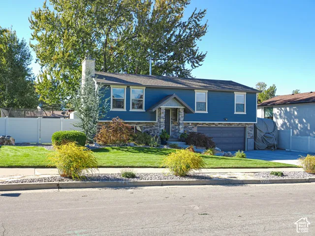 Split-level residence featuring blue siding, stone facade accents, an attached garage, a white picket fence, and a landscaped front yard with mature trees