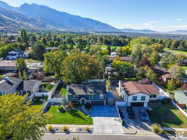 Striking blue contemporary home featuring a spacious driveway with multiple parking spaces, mature landscaping with established trees throughout the grounds, manicured lawn, and mountain backdrop