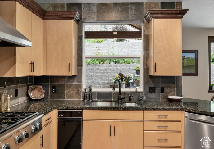 Light wood-finish cabinetry with dark hardware, stone tile backsplash, dark granite countertops, stainless steel undermount sink with a gooseneck faucet, and a stainless steel gas range