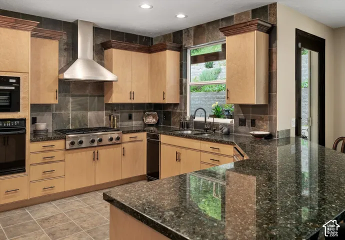 Kitchen featuring light wood cabinetry, stone tile backsplash, stainless steel range hood, built-in oven, and dark speckled countertops