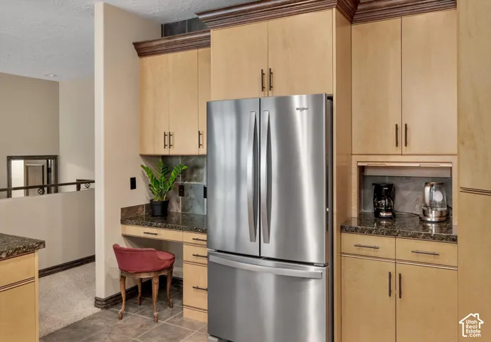 Light wood-finish cabinetry with dark hardware, stainless steel refrigerator, dark stone-finish countertops, tiled flooring, and a built-in desk area