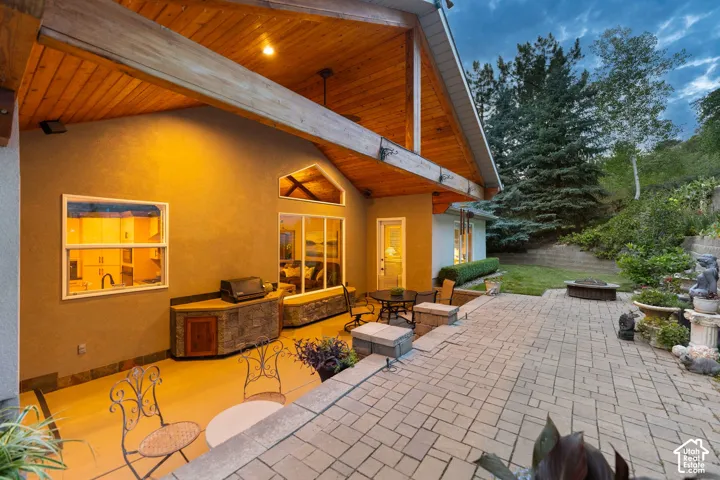 Expansive paver patio with a built-in outdoor kitchen, dedicated fire pit area, vaulted wood-paneled ceiling, and a sloped, landscaped yard