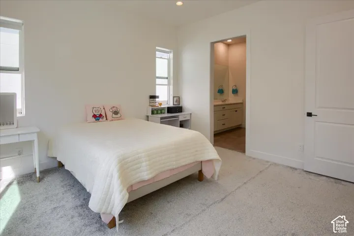 Recessed lighting illuminates the room, featuring two windows with roller shades, a panel door with black hardware, and an en-suite with a vanity and mirror