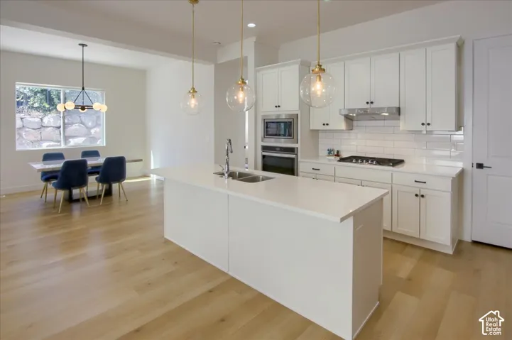 Expansive wood-finish flooring, white shaker cabinetry, stainless steel appliances, a large center island with integrated sink, and white subway tile backsplash