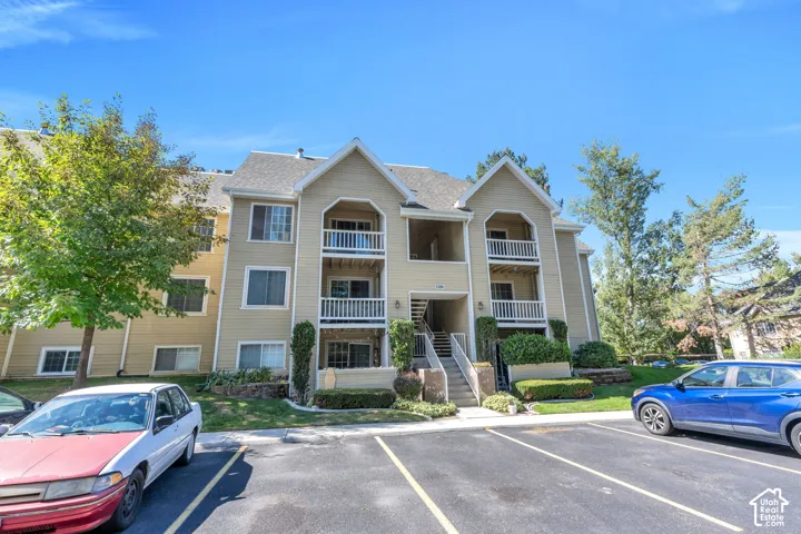 Multi-story residential building with light-colored siding, multiple balconies with white railings, a prominent central entryway staircase, and surrounding mature landscaping