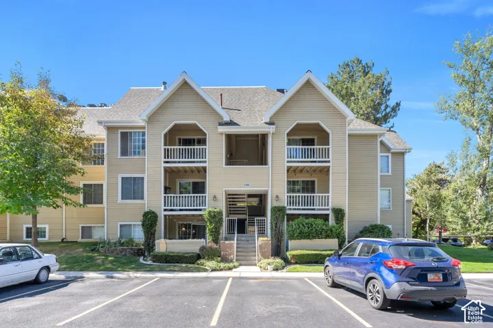 Multi-story building featuring balconies with white railings, a central entry staircase, and a parking area