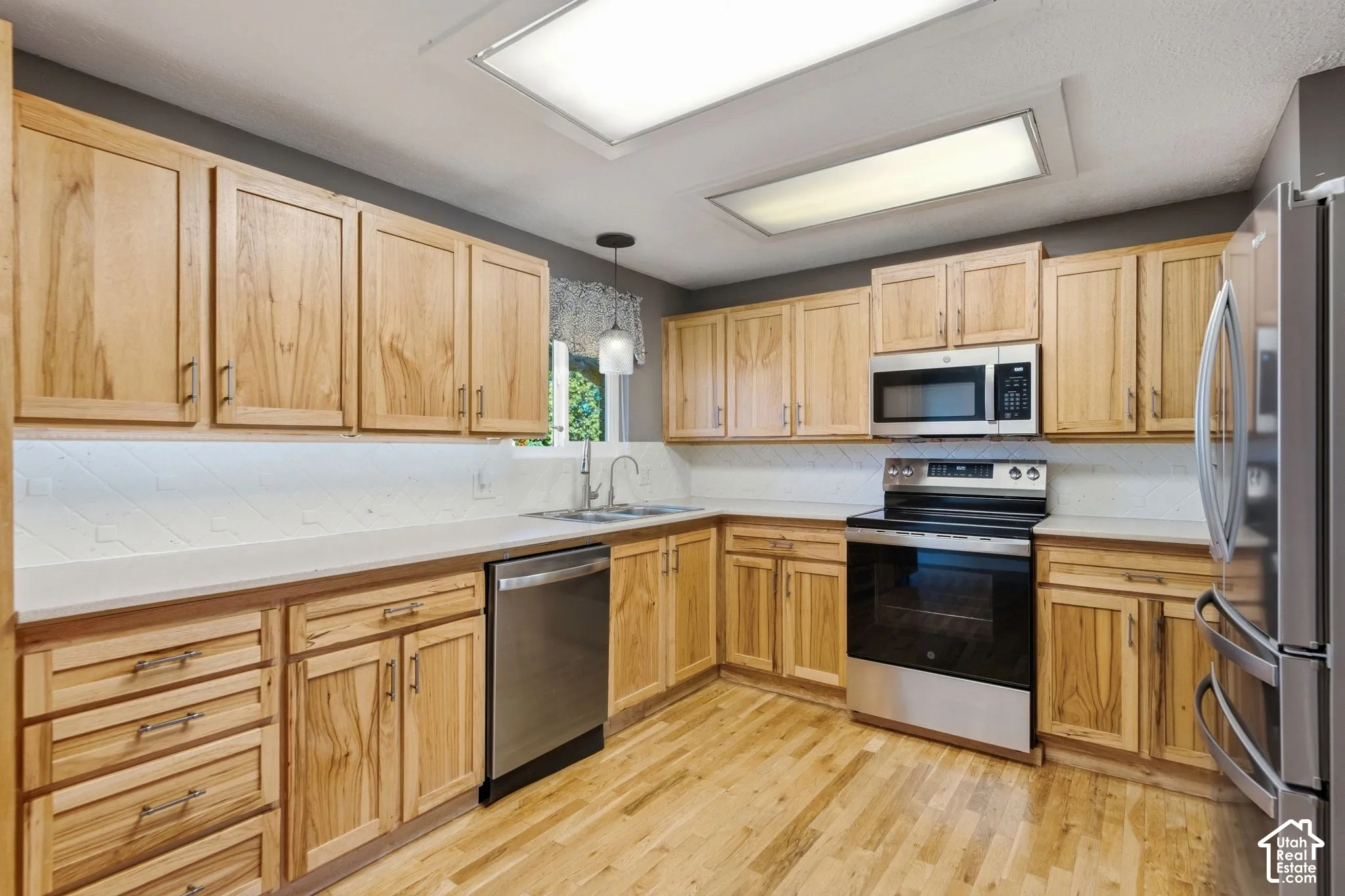 Kitchen with wood-finish cabinetry, stainless steel appliances, light-toned countertops, and a full tile backsplash