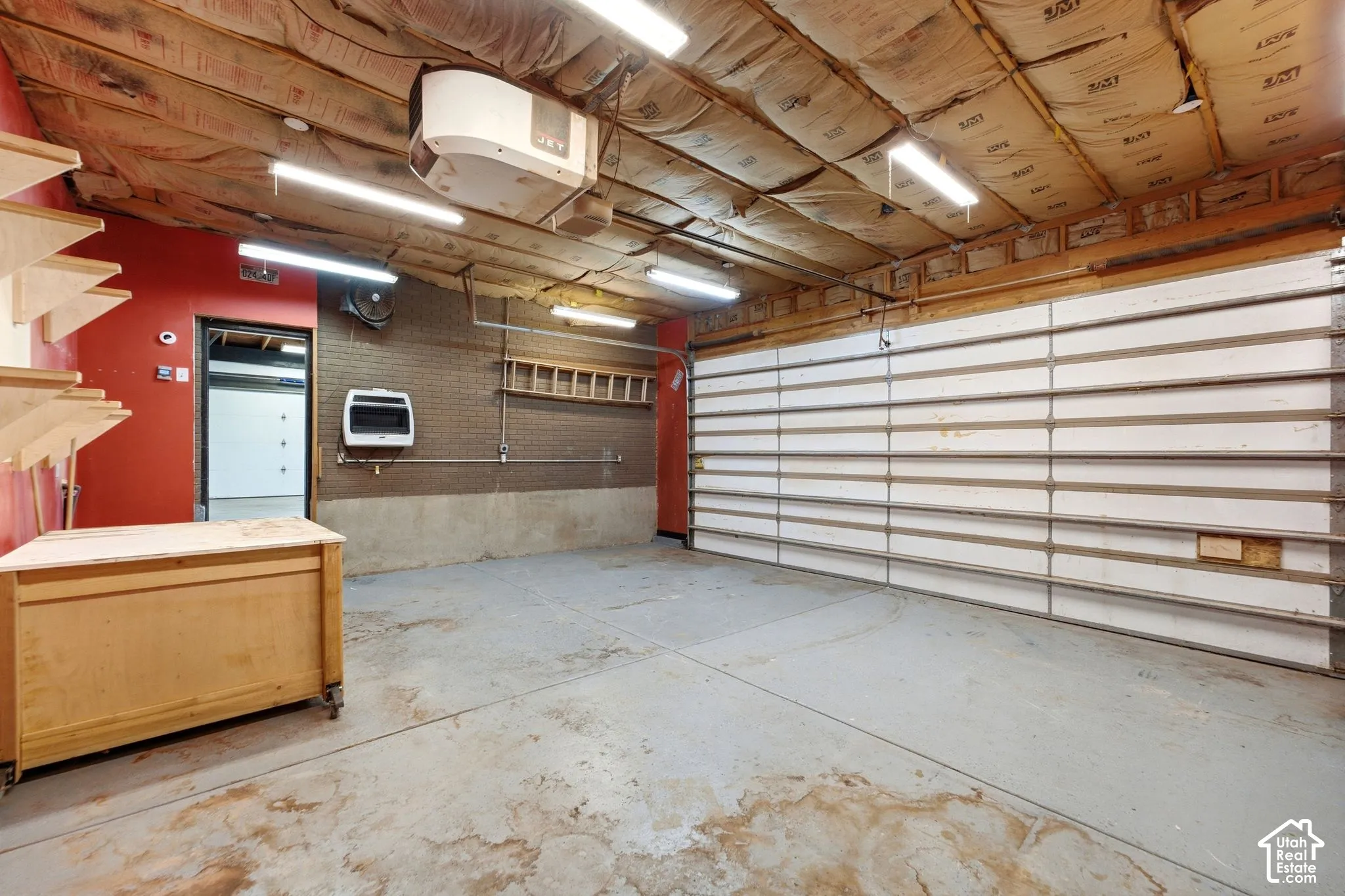 Concrete floor, exposed insulation ceiling, wall-mounted heater, track lighting, and a large garage door