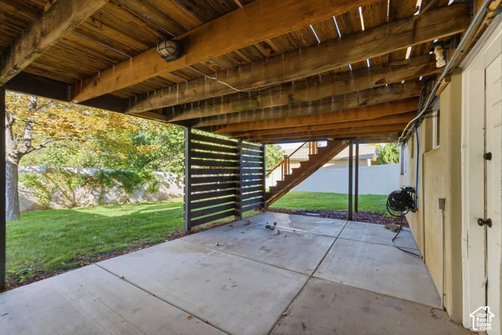 Concrete patio beneath a wood-beamed deck featuring a slatted wood privacy screen and exterior stairs