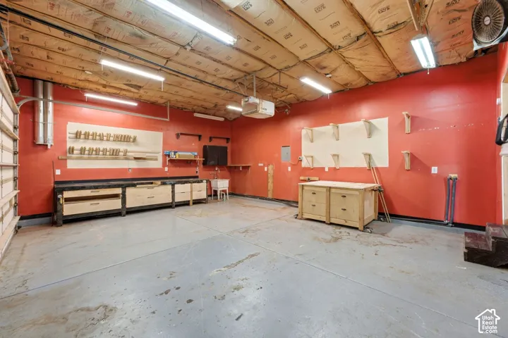 Spacious garage with exposed insulation ceiling, multiple fluorescent light fixtures, built-in workbench with drawers, and a utility sink