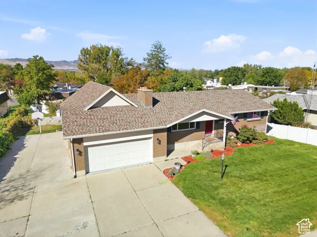Brick exterior with a shingled roof, extended driveway, attached garage, and manicured front lawn