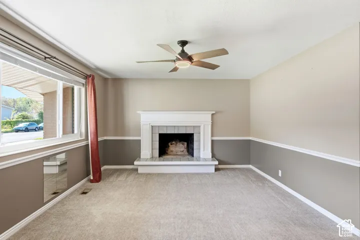 Carpeted room featuring a white mantel fireplace with tile surround, a ceiling fan with integrated lighting, and a large window
