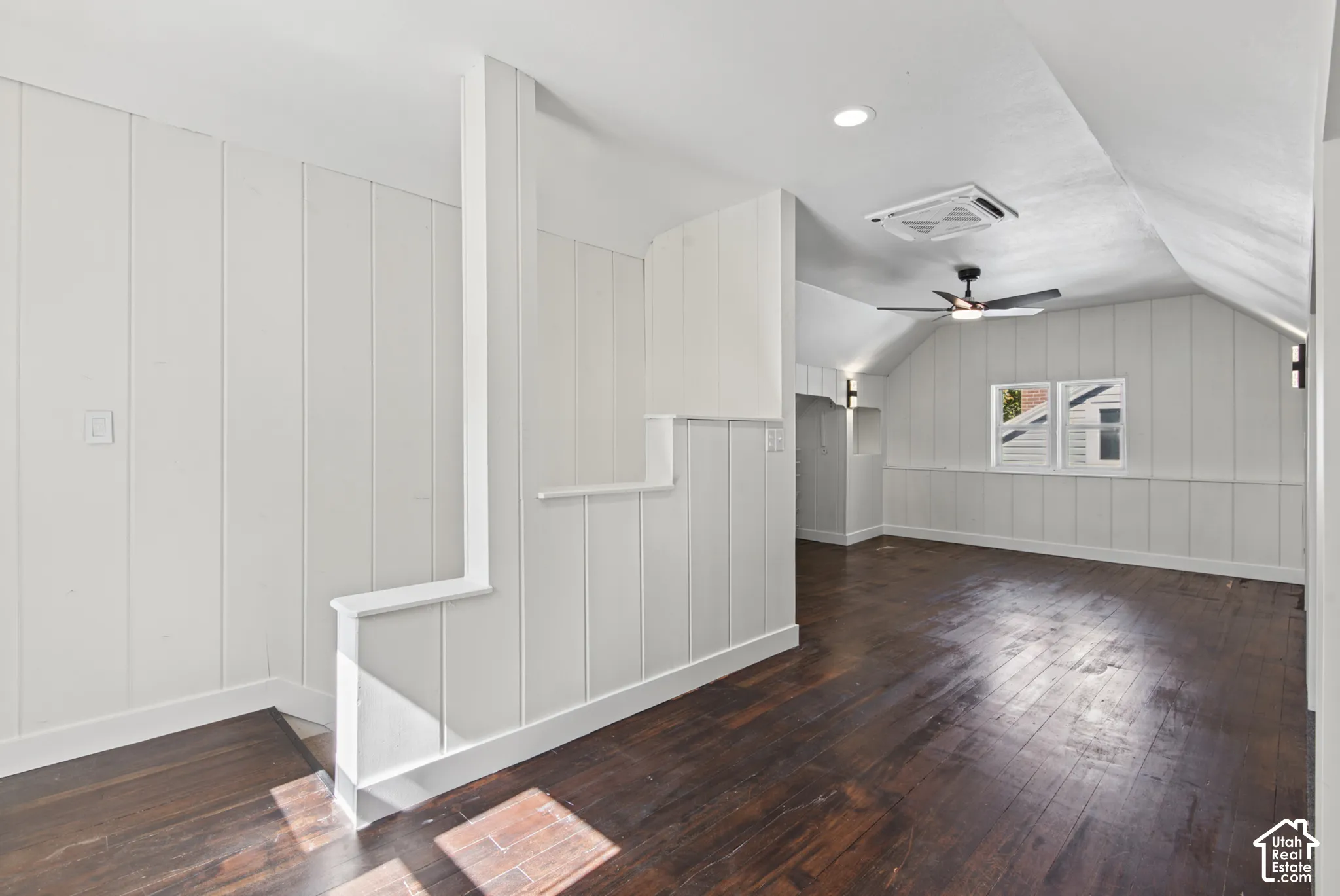 Dark wood-finish flooring, white vertical wall paneling, and a sloped ceiling with a ceiling fan