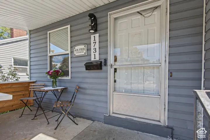Covered concrete porch with gray siding, featuring a white paneled entry door and house numbers