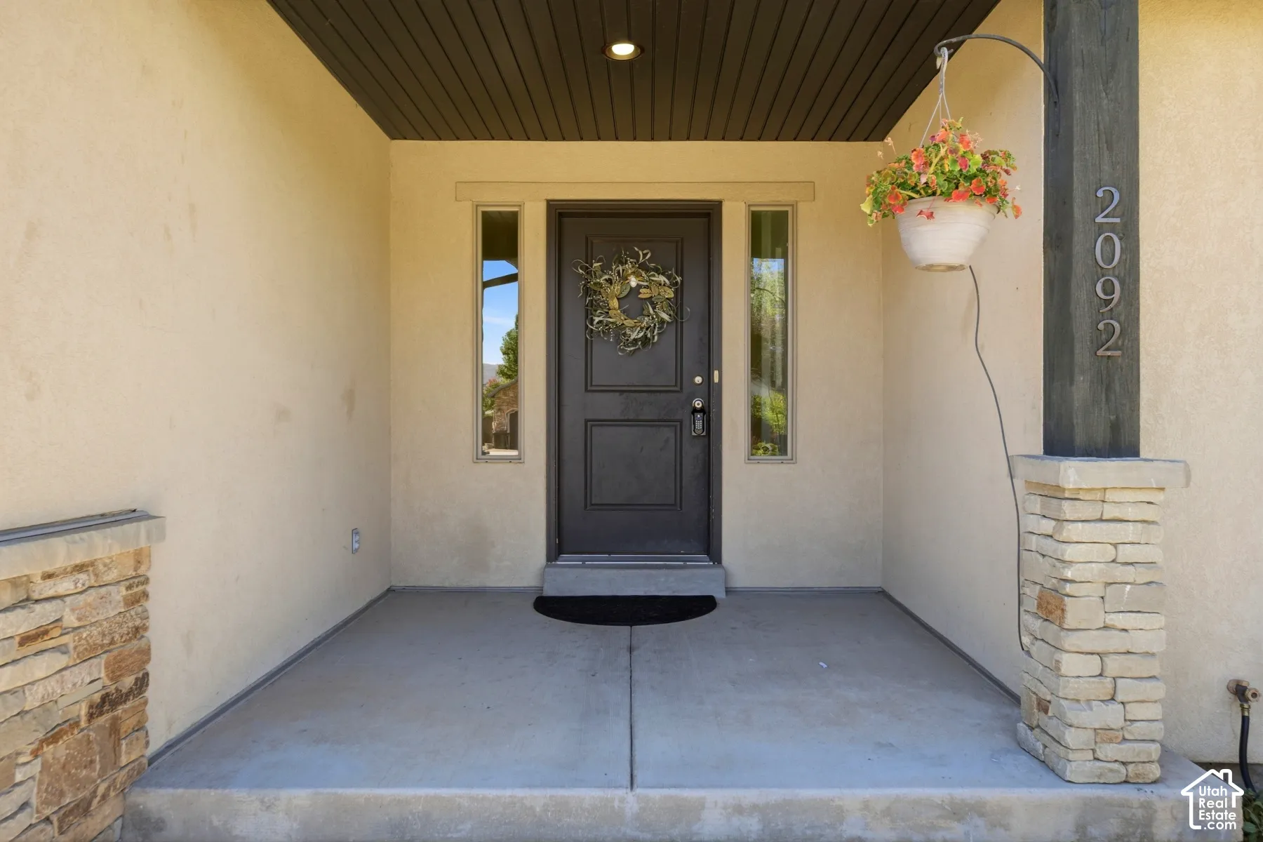 Covered front porch featuring a dark entry door with sidelight windows, stucco exterior, stone-clad pillar, and wood-finish ceiling
