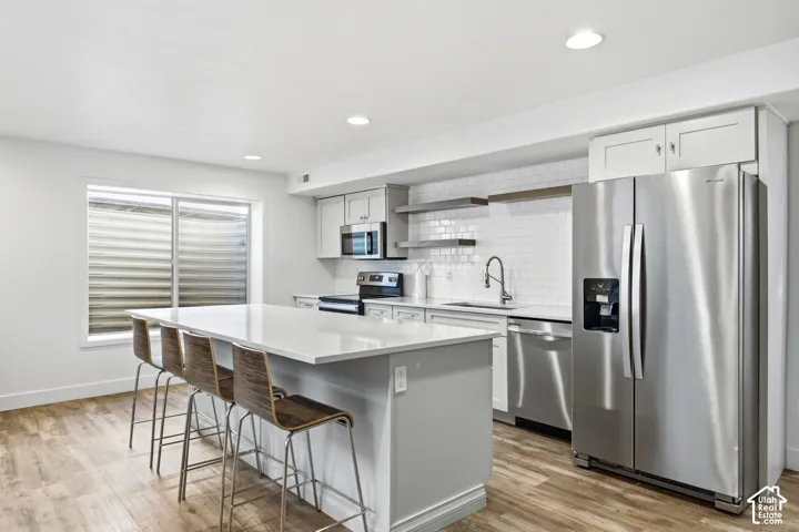 Open-concept kitchen featuring an oversized island, stainless steel appliances, light-toned cabinetry, subway tile backsplash, and wood-finish flooring