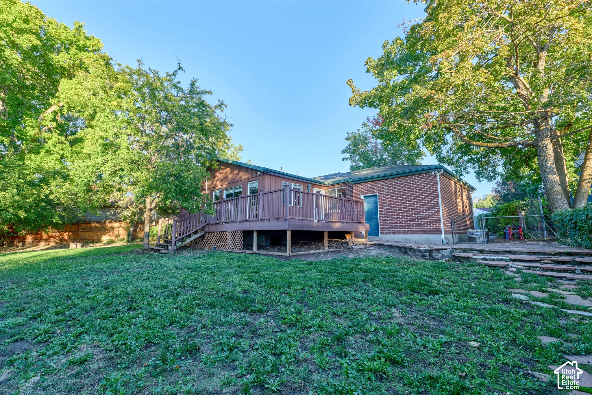 Expansive backyard featuring a large elevated wood deck with railing, exterior brick facade, and mature trees