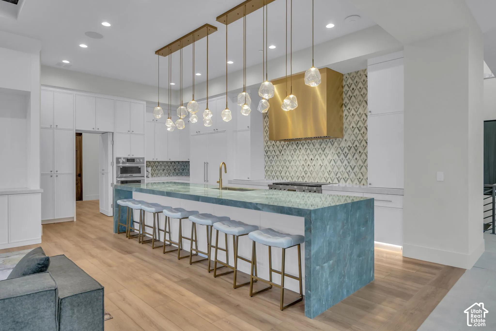 Extended kitchen island with a stone surface, brass pendant lighting, integrated wall oven, white cabinetry, and wood-finish flooring