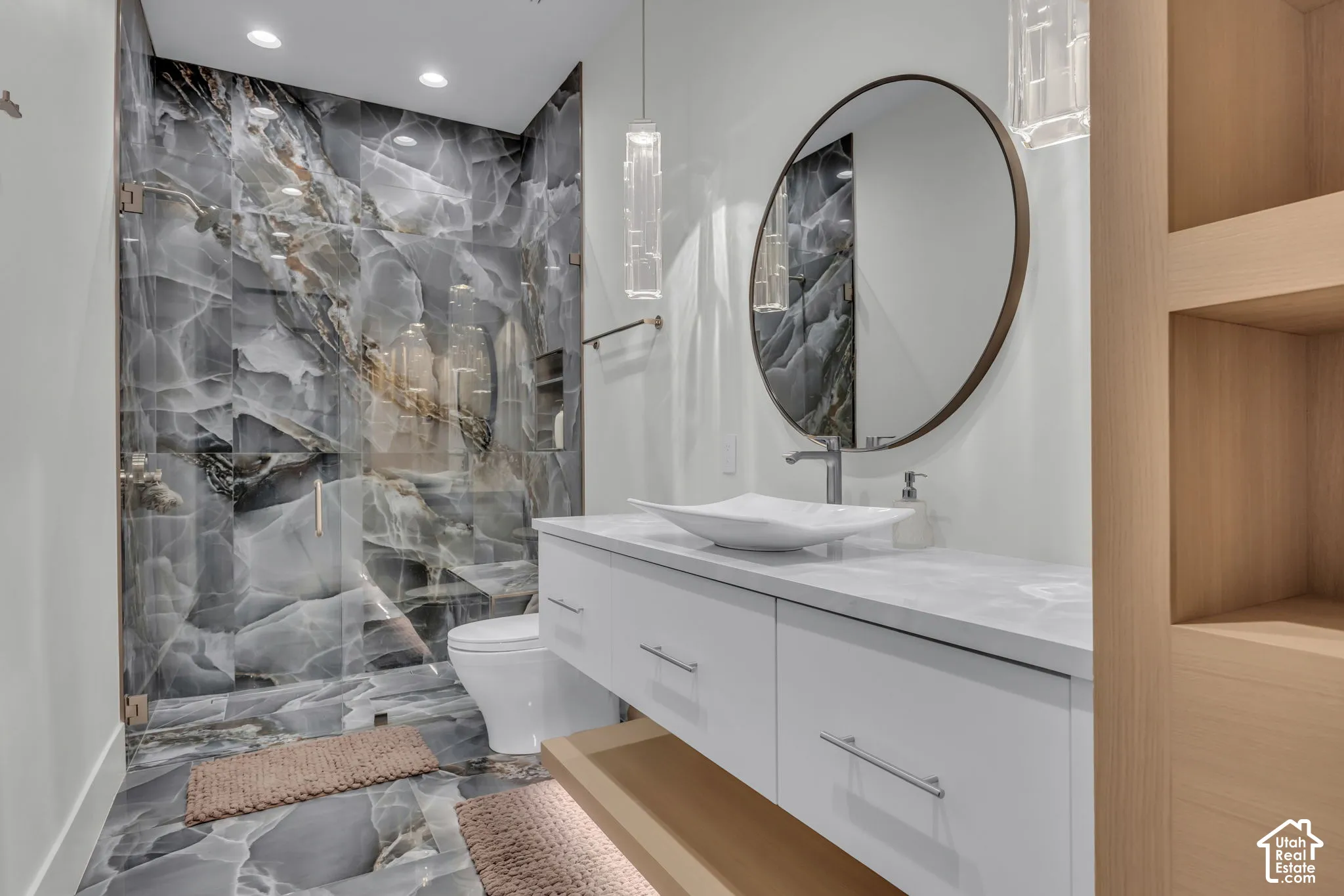 Glass-enclosed shower with large format stone-look tile, floating vanity with white vessel sink, rounded wall mirror, and integrated wood-finish shelving