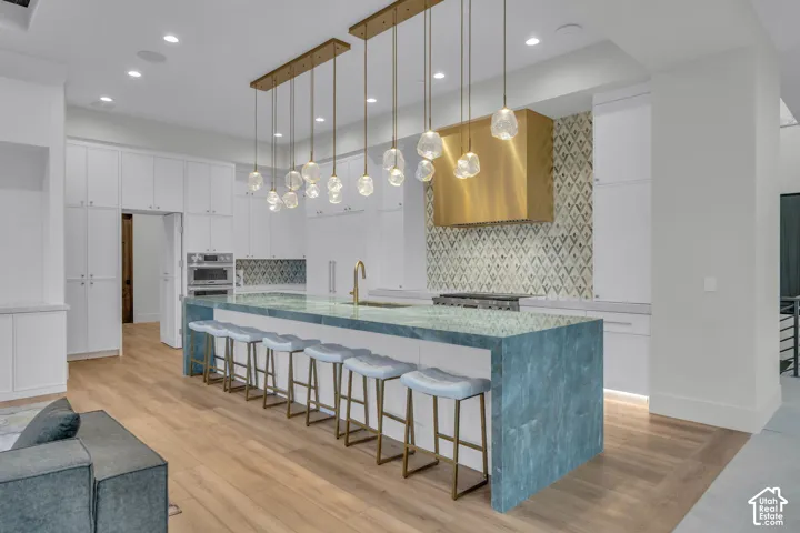 Extended kitchen island with a stone surface, brass pendant lighting, integrated wall oven, white cabinetry, and wood-finish flooring