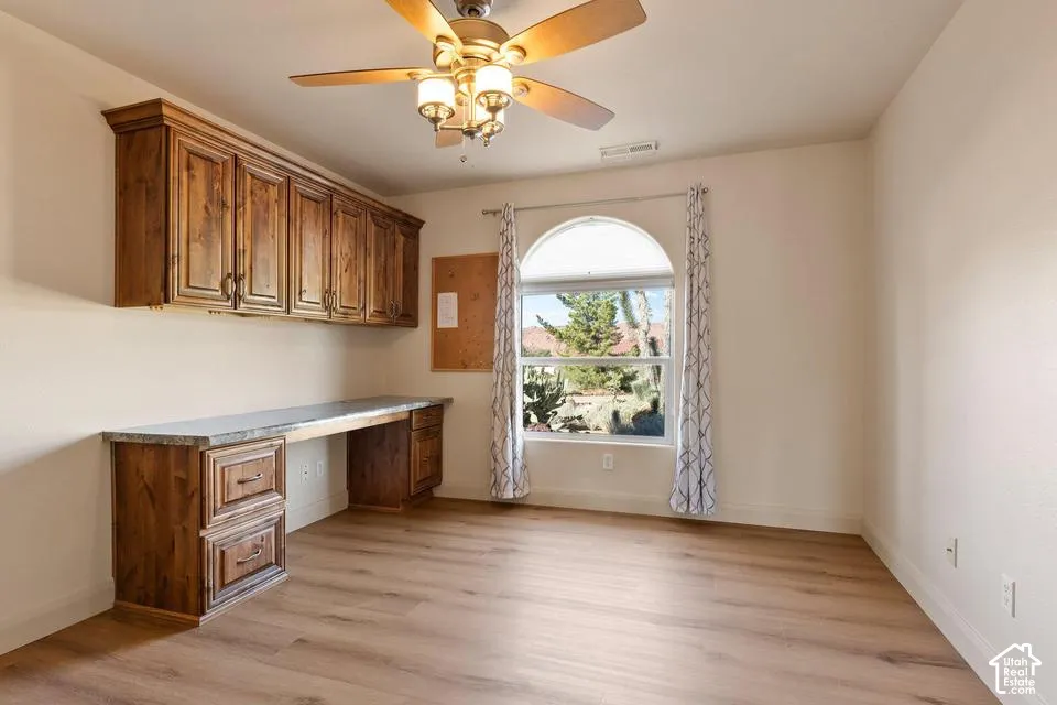 Built-in wood-finish cabinetry, wood-finish desk with a stone-finish countertop, wood-finish flooring, arched window, and ceiling fan with integrated lighting