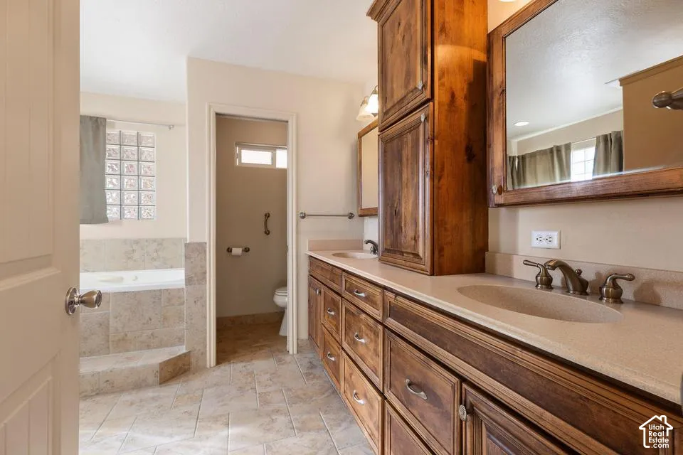 Double vanity with integrated sinks and bronze-finish fixtures, extensive wood cabinetry, built-in soaking tub with tile surround, and glass block privacy window