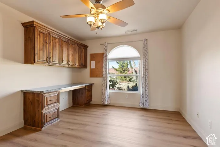 Built-in wood-finish cabinetry, wood-finish desk with a stone-finish countertop, wood-finish flooring, arched window, and ceiling fan with integrated lighting