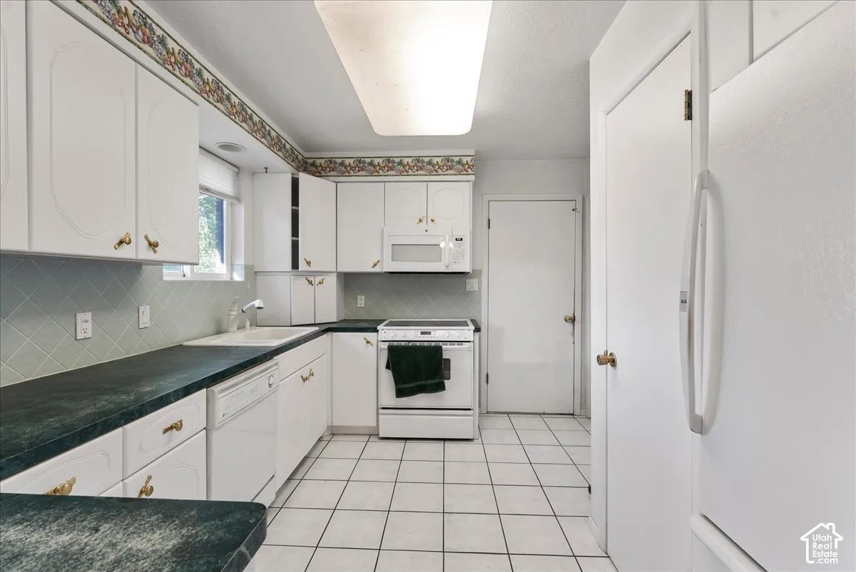 White cabinetry with brass-finish hardware, dark countertops, white tile flooring, and light green tiled backsplash