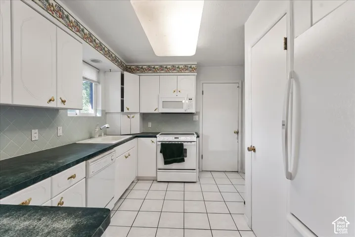 White cabinetry with brass-finish hardware, dark countertops, white tile flooring, and light green tiled backsplash