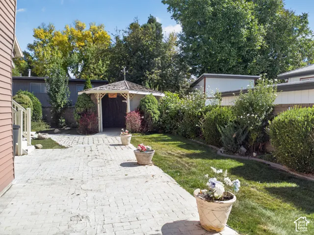 Stamped concrete patio with a detached gazebo, manicured shrubs, mature trees, and a grassy area