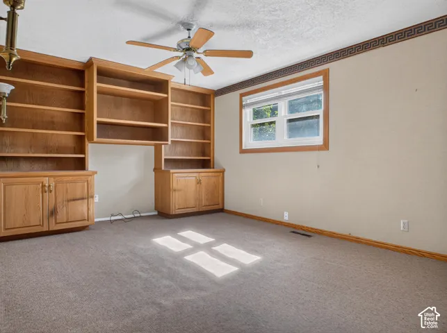 Built-in wood cabinetry with open shelving and closed storage, light brown carpeting, single window with wood trim, ceiling fan with light fixture, and decorative wall border