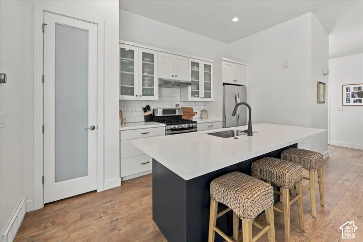 Kitchen featuring wood-finish flooring, white cabinetry with glass-front uppers, a kitchen island with seating, a stainless steel gas range, and a stainless steel refrigerator