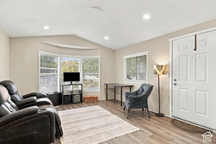 Vaulted ceiling with recessed lighting, wood-finish flooring, bay window with blinds, panel-style entry door, and a secondary window with blinds